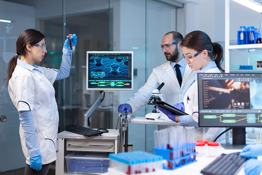 Team of medical research scientists conducting vaccine development with help of high tech, test tubes, micropipette and writing down analysis results on computer in modern equipped laboratory at night